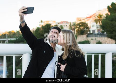 Gioiosa giovane coppia multirazziale in abbigliamento casual che porta selfie su telefono cellulare in piedi insieme su un ponte con alberi verdi e gli edifici della città sullo sfondo Foto Stock