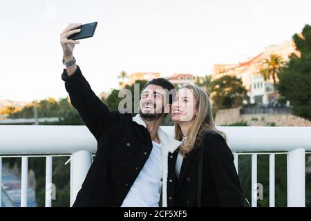 Gioiosa giovane coppia multirazziale in abbigliamento casual che porta selfie su telefono cellulare in piedi insieme su un ponte con alberi verdi e gli edifici della città sullo sfondo Foto Stock