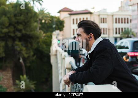 Vista laterale di un giovane maschio etnico in outfit alla moda sorridi e ascolta la musica con gli auricolari mentre ti riposi ponte della città Foto Stock