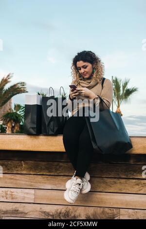 Soddisfatto giovane donna dai capelli ricci in abbigliamento casual sorridente mentre si utilizza il telefono cellulare su panca con borse della spesa sulla banchina moderna Foto Stock