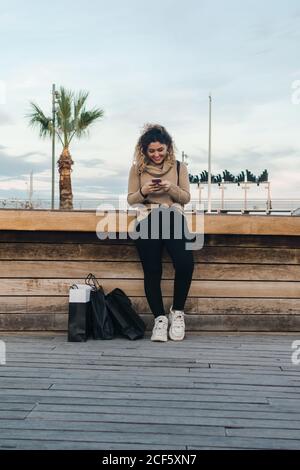 Soddisfatto giovane donna dai capelli ricci in abbigliamento casual sorridente mentre si utilizza il telefono cellulare su panca con borse della spesa sulla banchina moderna Foto Stock