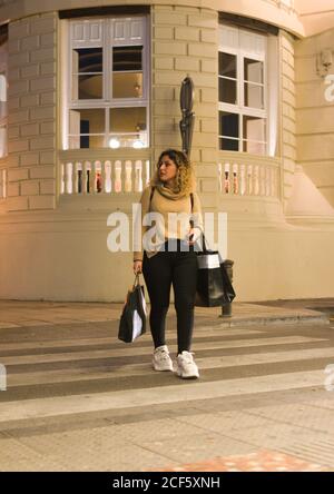 Giovane donna gioiosa in abbigliamento casual guardando lontano mentre attraversa una traversata zebra con telefono cellulare sulle mani e borse per lo shopping in città strada Foto Stock