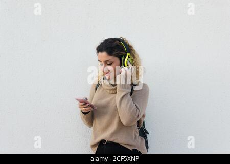 Giovane donna felice in beige maglione casual sorridente mentre si utilizzano cuffie e telefono cellulare con parete bianca sullo sfondo Foto Stock