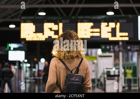 Vista sul retro di una donna anonima con zaino che guarda sopra imbarco con orario nel terminal dell'aeroporto Foto Stock