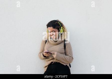 Giovane donna felice in beige maglione casual sorridente mentre si utilizzano cuffie e telefono cellulare con parete bianca sullo sfondo Foto Stock
