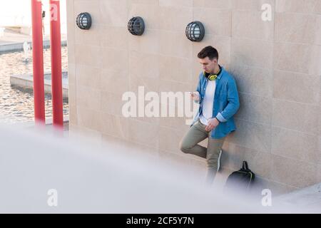 Uomo elegante e intelligente con cuffie brillanti che navigano sul telefono cellulare mentre appoggiandosi su un muro di marmo in una giornata di sole Foto Stock