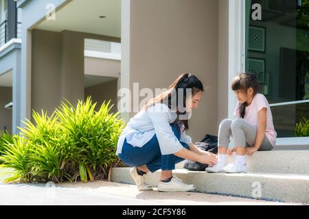 La madre asiatica aiuta la sua figlia studenti primari in uniforme a. indossando la loro scarpa davanti a casa nel la routine scolastica mattutina per il giorno in Foto Stock