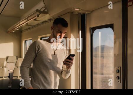 Giovane uomo concentrato in wireless auricolari messaggi di testo sul cellulare in piedi e appoggiati alla parete del treno alla luce del sole Foto Stock
