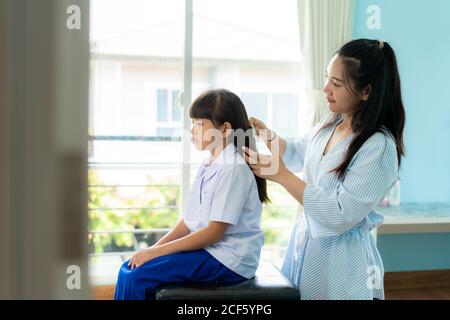 La madre asiatica sta combattendo i capelli della figlia la mattina prima di andare a scuola nel soggiorno a casa. La routine scolastica mattutina per il giorno negli li Foto Stock