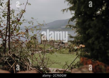 Moody Scottish paesaggio con rovine di vecchio castello situato su riva del lago contro le colline foggose incorniciate da rami di alberi Foto Stock