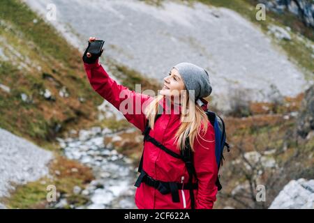 Dall'alto sorridente Donna turista in giacca rossa con zaino prendere selfie su telefono cellulare sullo sfondo di verdi montagne rocciose in villaggio nelle vette d'Europa, Asturie, Spagna Foto Stock