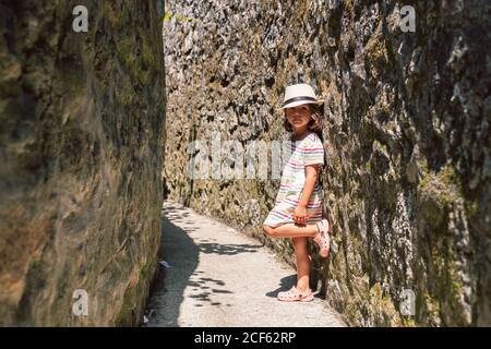 Bambino adorabile attivo in cappello appoggiato su parete di pietra dentro luce solare e guardando la fotocamera Foto Stock