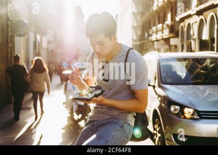 Bell'uomo etnico asiatico che mangia cibo da asporto mentre si sta in piedi camion di cibo sulla strada soleggiata Foto Stock