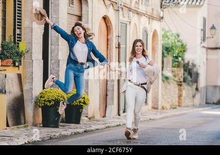 Giovani donne sorridenti che saltano e si divertono a tenere le mani saltando in una bella strada decorata con fiori Foto Stock