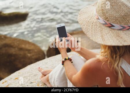 Donna moderna in cappello di paglia seduto sulla pietra costiera e tenendo smartphone durante le vacanze Foto Stock