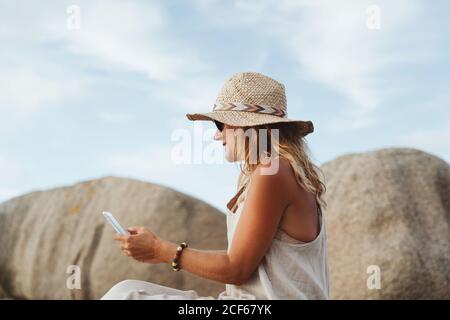 Donna moderna in cappello di paglia seduto sulla pietra costiera e tenendo smartphone durante le vacanze Foto Stock