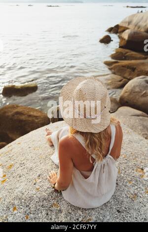 Vista posteriore della donna felice e ottimista in cappello di paglia seduto di fronte alle scogliere di riva e sorridente dentro luce del tramonto Foto Stock