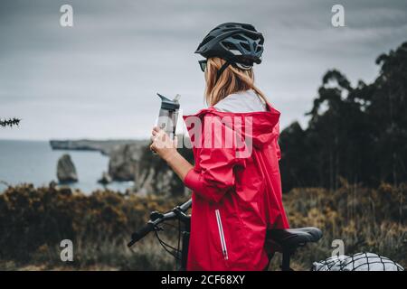 Vista laterale della donna in bicicletta con casco e giacca rossa bere acqua e godere di un paesaggio pittoresco Foto Stock