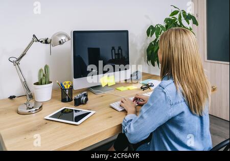 Vista posteriore o bionda donna caucasica in abiti casual seduto su una scrivania di legno lavorando su un computer in un moderno ufficio domestico Foto Stock