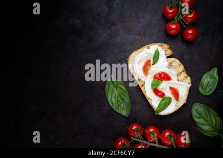 Vista dall'alto del brindisi croccante cosparso di formaggio cremoso e. decorato con foglie di basilico e pezzi di pomodori ciliegini sfondo nero Foto Stock