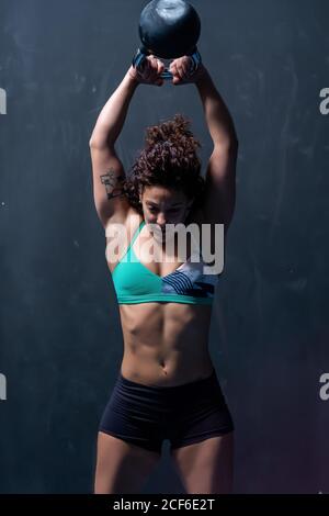 Potente sportswoman in reggiseno e pantaloncini che sollevano il kettlebell pesante nelle braccia sollevate sopra la testa durante gli allenamenti intensi di sollevamento pesi in palestra Foto Stock