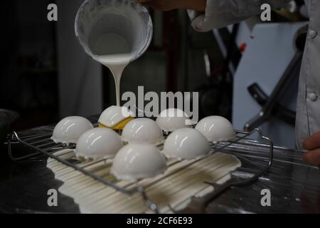 Crop persona versando bianco specchio glassa su gustosa mousse gialla torte disposte su griglia metallica in cucina Foto Stock