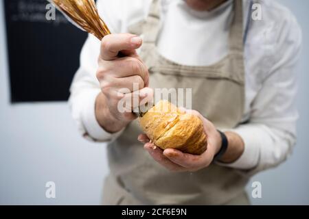 Coltivare l'uomo in grembiule e ripieno bianco uniforme croissant fresco con crema al cioccolato all'interno Foto Stock