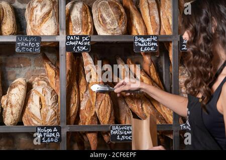 Donna irriconoscibile in grembiule nero mettendo baguette francese in sacchetto di carta da thongs di metallo in panetteria Foto Stock