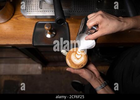 Dall'alto, le mani di un dipendente professionista preparano il cappuccino con in cima alla caffetteria Foto Stock