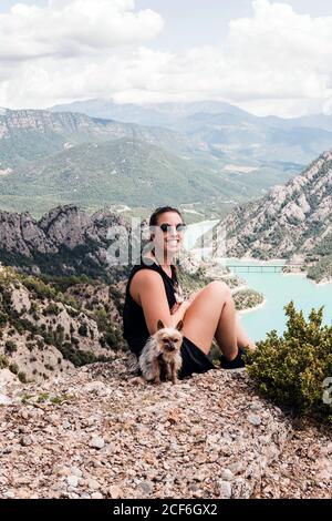 donna turistica in abiti neri casual e occhiali da sole seduti con dog su una collina rocciosa e ammirando paesaggi pittoreschi in estate Foto Stock