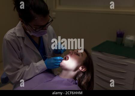 Stomatologo femminile in maschera e occhiali che esaminano i denti della giovane Donna con gli strumenti dentisti in clinica stomatologica Foto Stock