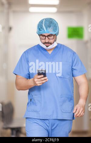 Chirurgo maschile in piedi nel corridoio durante il controllo dei messaggi il suo smartphone Foto Stock