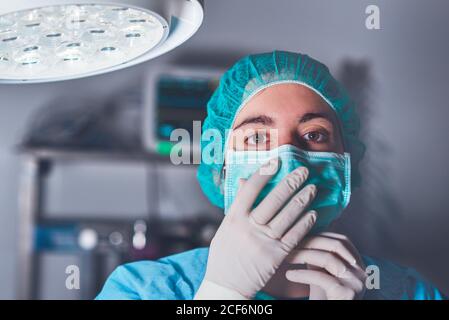 Donna adulta in maschera medica e cappello guardando la macchina fotografica prima di eseguire un intervento chirurgico in ospedale Foto Stock