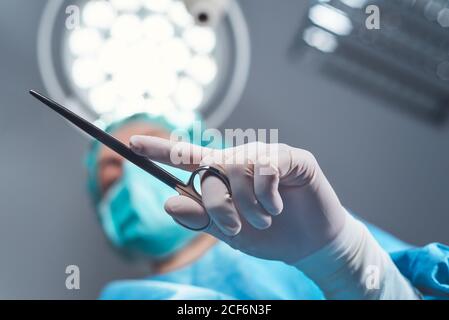 Da sotto chirurgo donna in uniforme medica utilizzando strumenti professionali in piedi sotto la luce luminosa in sala operatoria Foto Stock