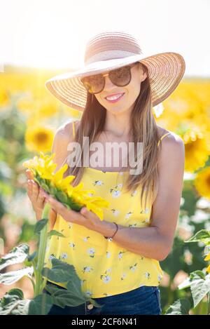 Donna in cappello di paglia che guarda mentre si sta in piedi su girasole luminoso campo Foto Stock