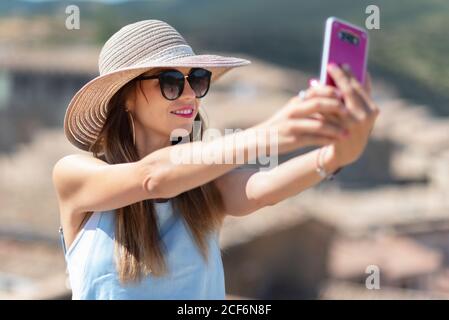 Donna spensierata in cappello e occhiali da sole che scattano foto in smartphone e sorridono mentre si trova in piedi per strada nella città panoramica Foto Stock