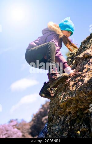 Vista laterale della ragazza carina in abiti caldi che arrampicano ruvido scogliera rocciosa contro il cielo blu nella giornata di sole in campagna Foto Stock