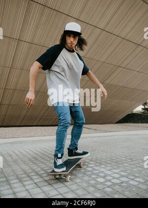 Un ragazzo millenario in equilibrio dei cappelli mentre cavalcano lo skateboard strada della città sotto un muro inclinato Foto Stock