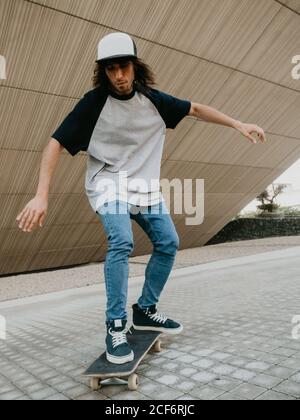 Un ragazzo millenario in equilibrio dei cappelli mentre cavalcano lo skateboard strada della città sotto un muro inclinato Foto Stock