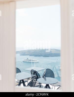 Vista pittoresca di una piccola terrazza con ringhiere in vetro e nero tavoli e sedie che si localizzano sullo sfondo di un meraviglioso mare blu con nave da crociera e cielo limpido Foto Stock