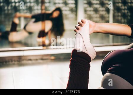 Ritaglio di ballerina che tira la gamba per allungare i muscoli seduti sul pavimento in studio di balletto. Foto Stock