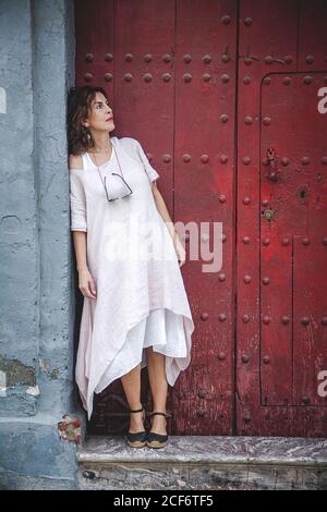 Pensieroso serio sognando donna elegante in abito estivo pendente sul muro vicino all'antica porta di maroon in città che guarda lontano Foto Stock