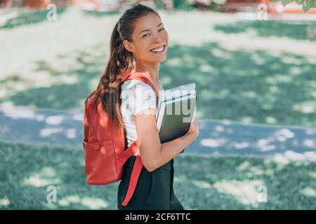 Felice studente universitario. Giovane donna asiatica sorridente alla macchina fotografica, camminando nel campus con zaino, laptop, libro. Concetto di ritorno a scuola Foto Stock