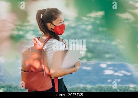 Torna alla scuola studentessa asiatica donna indossando maschera sul campus camminando con zaino rosso, libri e laptop. Coronavirus realtà Foto Stock