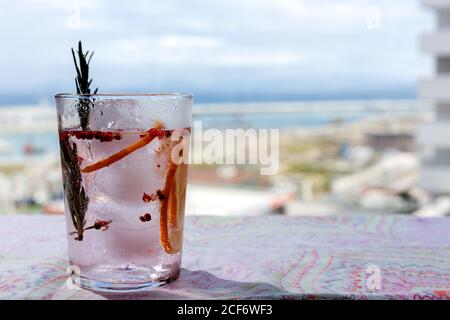 Cocktail tonico gin con acqua tonica rosa, pepe rosa, rosmarino, menta, cannella, limone e arancia al sole su un tavolo da ristorante Foto Stock