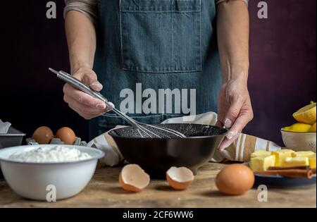 Crop anonymous Woman in grembiule montare le uova in ciotola nera su tavola di legno con limone, farina, burro e cannella ingredienti per la torta Foto Stock