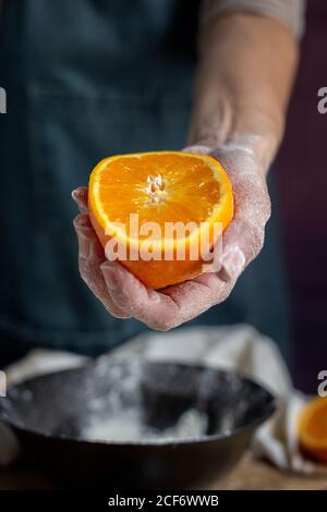 Ritaglia la mano di Donna irriconoscibile coperta di farina che tiene e mostra alla fotocamera un fresco mezzo taglio di arancia sul recipiente mentre prepari l'impasto a tavola Foto Stock