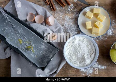 Vista dall'alto degli ingredienti per la ricetta della torta, inclusa la ciotola con farina e uovo posti su tavola di legno spolverato Foto Stock