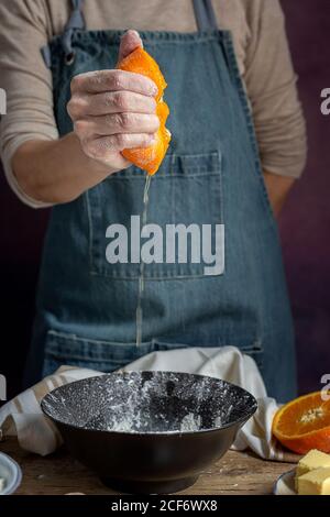 Ritaglia la mano di donna irriconoscibile in grembiule spremendo fresco succosa arancia tagliata sul recipiente mentre prepari l'impasto a tavola Foto Stock