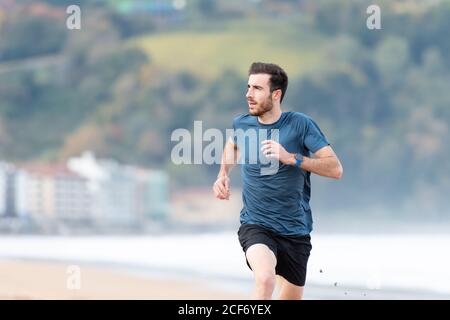 Atleta maschile sopportato in usura attiva che corre durante la sabbia vuota spiaggia con montagne verdi su sfondo sfocato Foto Stock